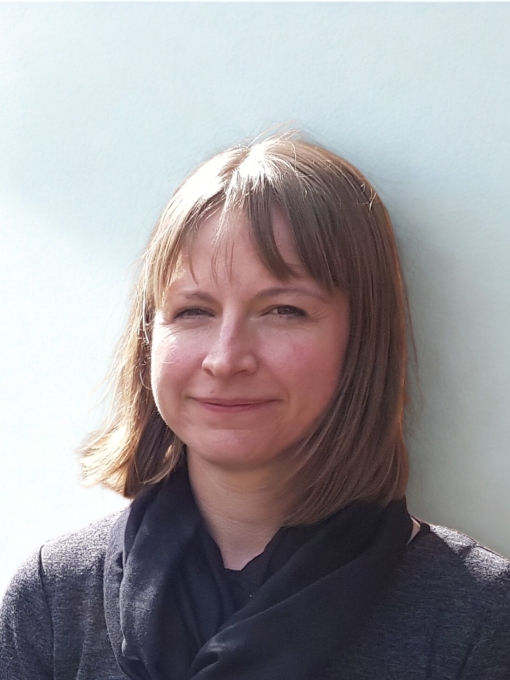 A profile photo of a woman wearing a dark coat and scarf with shoulder length brown hair. 