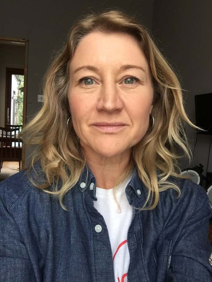 Woman with medium length wavy blonde hair in a denim shirt, sitting indoors with a doorway in the background.