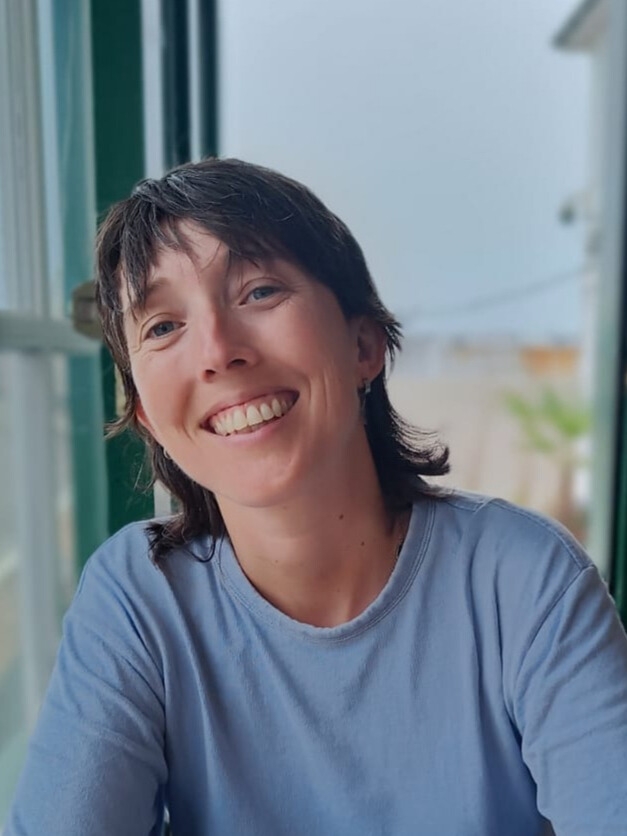 Woman with shoulder-length hair in a light blue shirt sitting indoors near a window.