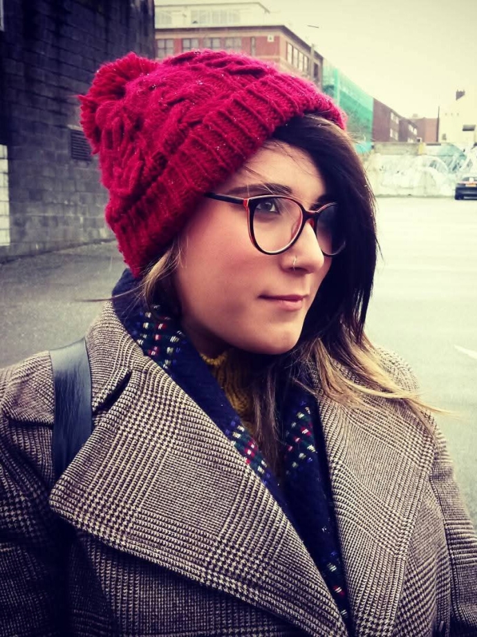 Woman in red beanie, glasses, checkered coat, and scarf standing on a city street.