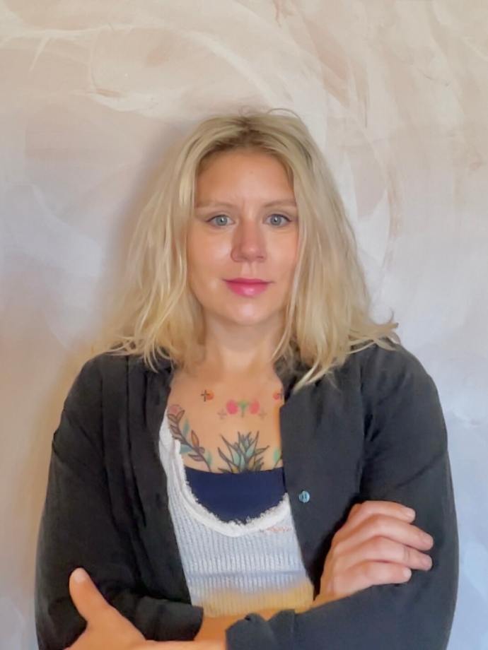 Woman with blonde medium length hair, wearing a dark cardigan and white tank top, standing with arms crossed.