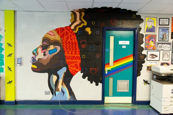 A school hallway with a vibrant mural of a black woman's head and hair is painted across the whole wall and over a doorway.
