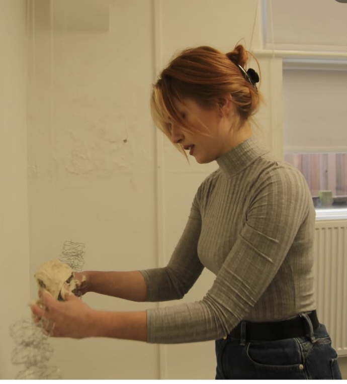 A person with reddish-brown hair tied up, wearing a gray ribbed turtleneck and dark jeans, is arranging a light-coloured object and wire structures on a white wall in a bright indoor space.