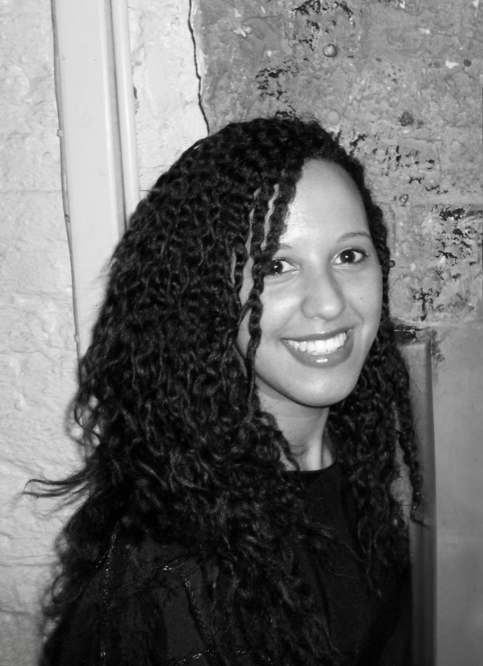 A black-and-white photo showing a person with long, voluminous, tightly curled hair wearing a dark top. The background features a textured wall with peeling paint and a vertical pipe running along the left side.