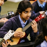 Girl sitting on the floor of a classroom playing a ukulele.