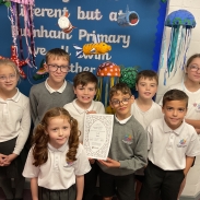 Group of schoolchildren standing in front of a colourful underwater-themed display board decorated with jellyfish, fish, and sea creatures. The board includes text that reads “Different but at Burnham Primary we all swim together.” One child in the front holds an Artsmark certificate or artwork with a decorative border.