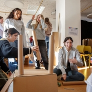 Group of people engaged in a hands-on creative activity indoors, building structures with cardboard tubes and blue blocks, with others standing and observing in the background.